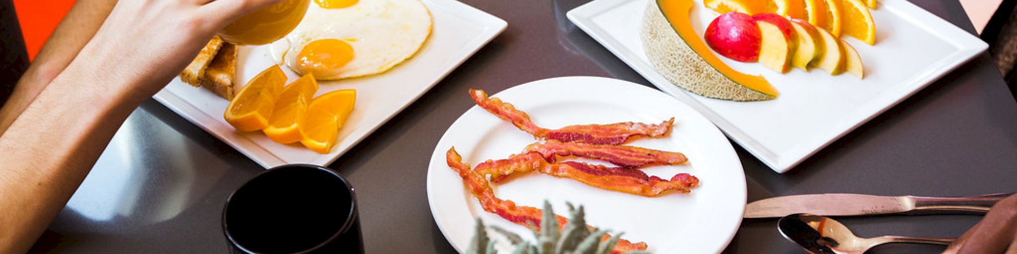 Two people dining with plates of eggs, bacon, fruits, a cup of coffee, and a glass of juice on a table.