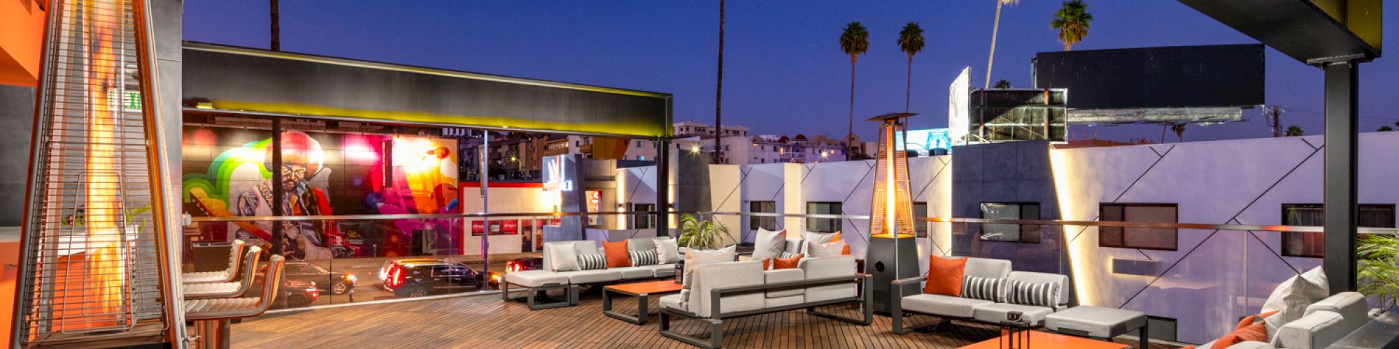 An outdoor lounge with sofas, heaters, and palm trees at dusk.