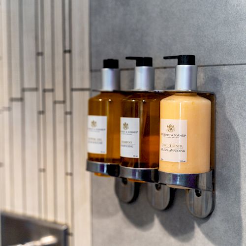 The image shows three bottles of liquid soap or shampoo mounted in a bathroom dispenser on a tiled wall.