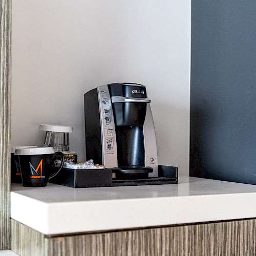 A coffee maker sits on a counter with a black mug and a small tray of coffee pods and condiments next to it, against a white and dark wall.