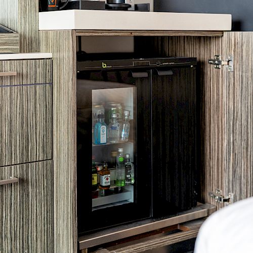 The image shows an open cabinet with a mini-fridge inside, containing various small bottles and beverages. The cabinet is next to a bed.