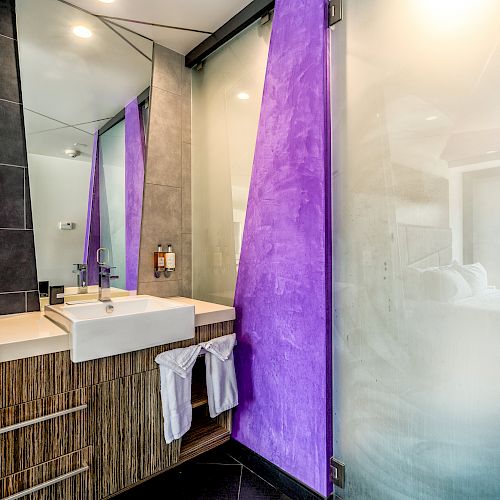 A modern bathroom with a glass door shower, a sink with a large mirror, and purple accents on the walls. Towels are hanging on the cabinet.