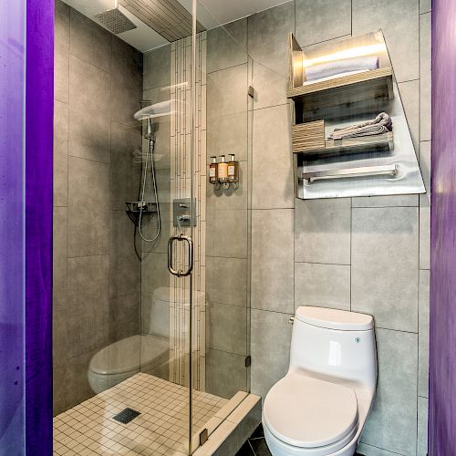 A modern bathroom with a glass-enclosed shower, gray tiles, a toilet, and a wall-mounted shelf holding towels and toiletries, with purple walls.