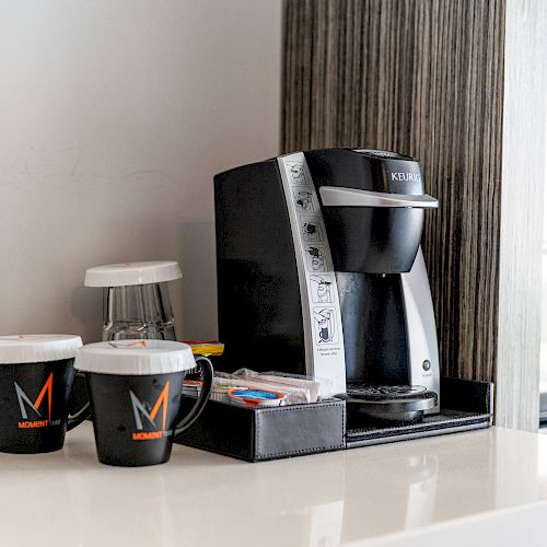 A coffee maker on a counter with two branded coffee mugs, a small tray containing coffee pods, and some complimentary items.