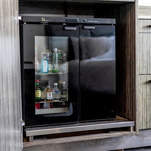 The image shows a small glass-door refrigerator built into a wooden cabinet, containing several bottles of various beverages.