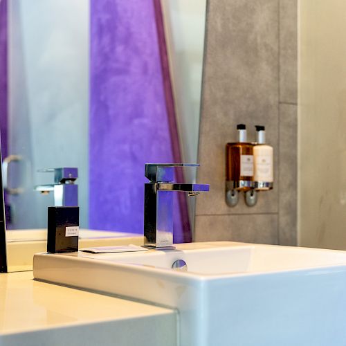 A modern bathroom with a sleek sink, minimalist chrome faucet, and wall-mounted soap dispensers. The decor includes purple and gray accents, ending the sentence.