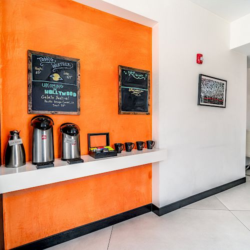 A coffee station with thermoses, cups, and chalkboard menus on an orange wall, in a modern, minimalistic room.