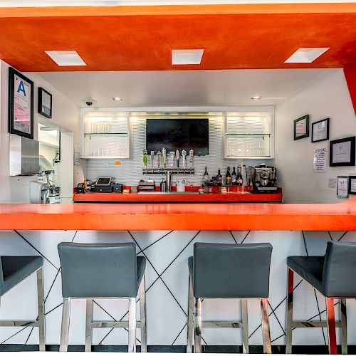 A modern bar with a bright orange counter, four gray stools, a TV, bottles, and framed pictures on the wall is shown in the image.