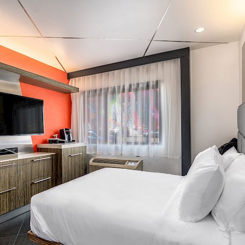 A modern hotel room features a bed, flat-screen TV, kitchenette with a coffee maker, and large window with curtains.