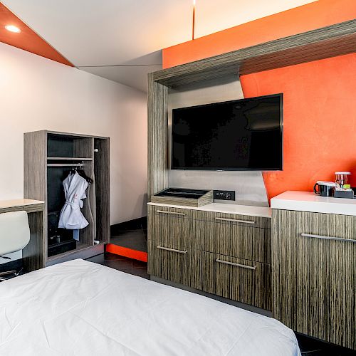 A hotel room features a bed, desk with chair, open wardrobe, wall-mounted TV, and a cabinet with a coffee maker on an orange and white color scheme wall.