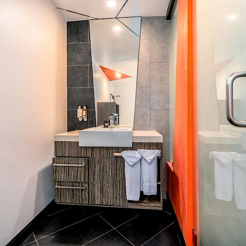 A modern bathroom with a sink, a mirror, a wooden vanity cabinet, neatly hanging towels, and a shower area with a glass door ending the sentence.