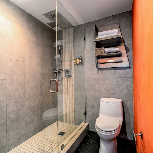 The image shows a modern bathroom with a glass-enclosed shower, a toilet, gray tiles, and a wall-mounted shelf holding towels and toiletries.