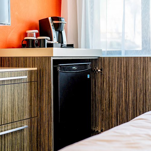 The image shows a modern hotel room setup with a coffee maker, mini-fridge, and wooden cabinets next to a bed with white linens ending the sentence.