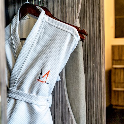 A white bathrobe with an embroidered "M" logo is hanging on a wooden hanger in a modern bathroom setting with wooden cabinetry.