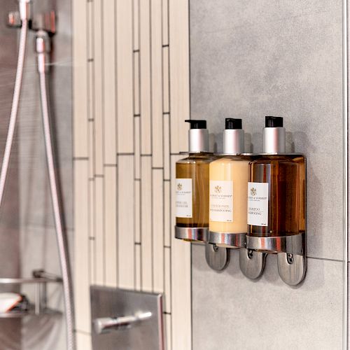 The image shows a modern shower setup with three wall-mounted dispensers containing shampoo, conditioner, and body wash, next to a showerhead.