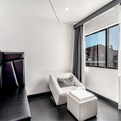 A minimalist room featuring a black padded bench, a white armchair with a cushion, an ottoman, a large window with curtains, and white walls.