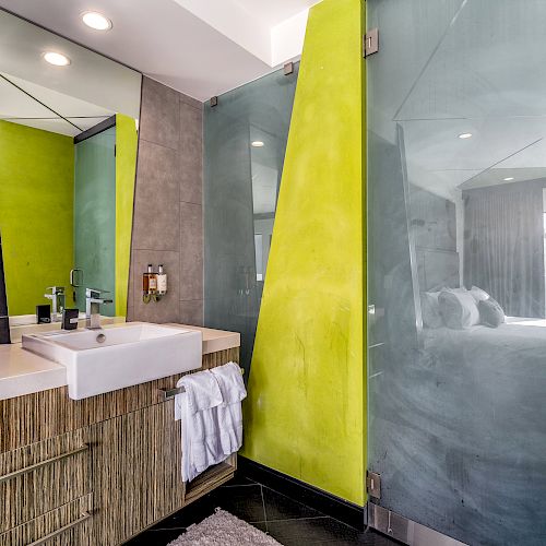 A modern bathroom with a green and black color scheme, featuring a sink, mirror, towels, and a walk-in shower with glass doors, leading to a bedroom.