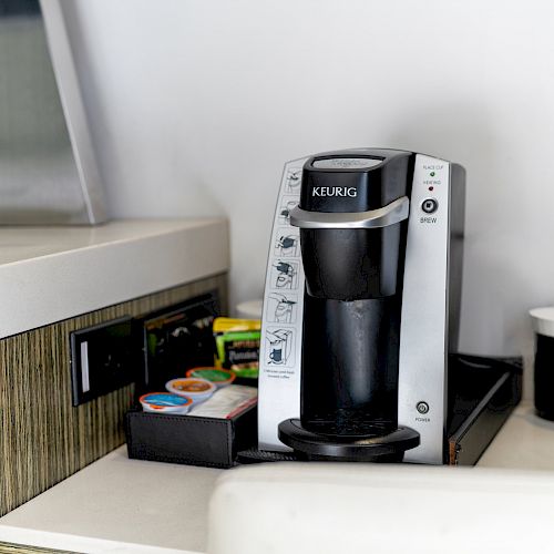 A Keurig coffee machine is on a counter, next to a black coffee mug and a tray containing various coffee pods.