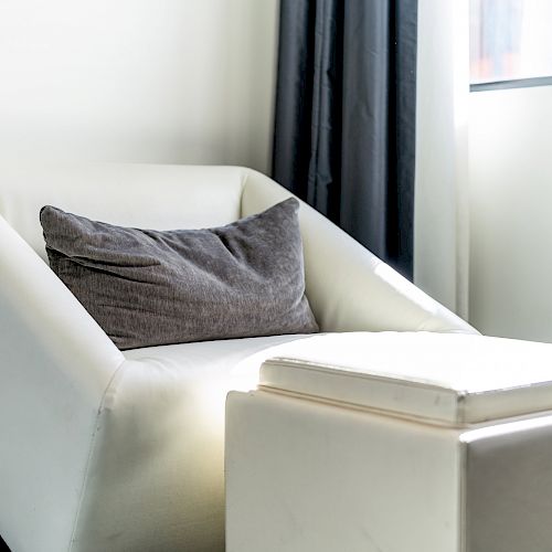 The image shows a modern white armchair with a gray cushion beside a white ottoman, near a window with black curtains.