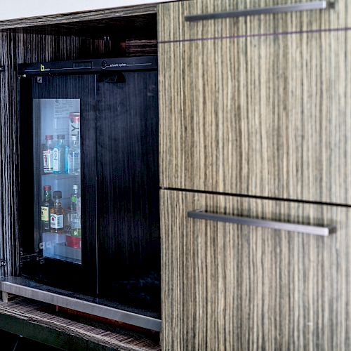 The image shows a wooden cabinet with an open door, revealing a mini fridge stocked with various small beverage bottles inside.