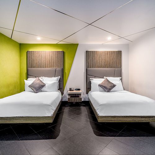 The image shows a modern hotel room with two single beds, green and white walls, and a central nightstand between the beds, ending the sentence.