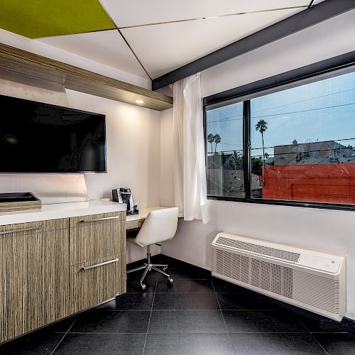 The image shows a modern room with a wall-mounted TV, a desk with a white chair, a large window, and air conditioning.