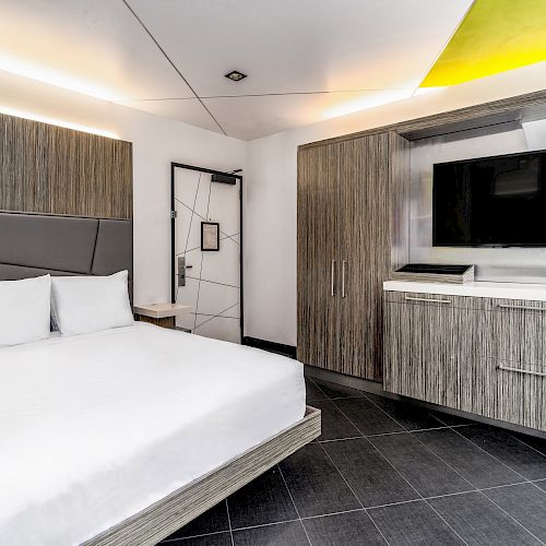 A modern hotel room features a bed, TV, cabinets, and a workspace with a chair. The decor is sleek with white and wooden tones, and ample lighting.