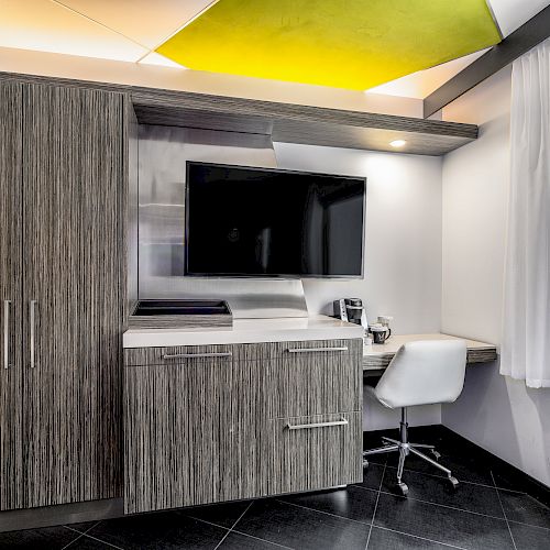 A sleek modern workspace with a wall-mounted TV, wooden cabinets, a white chair, desk, and a window with white curtains.