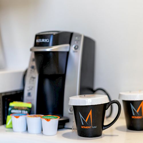 A Keurig coffee maker, several coffee pods, and two mugs with "M" logos are placed on a countertop.