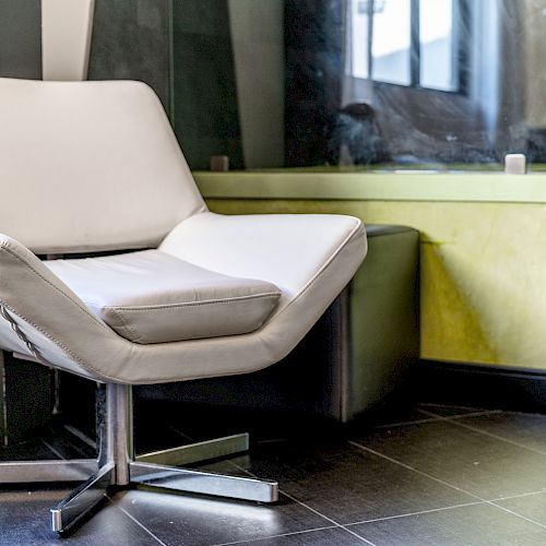 A modern white armchair with a metal base is placed beside a yellow cabinet on a tiled floor in a room with large windows.