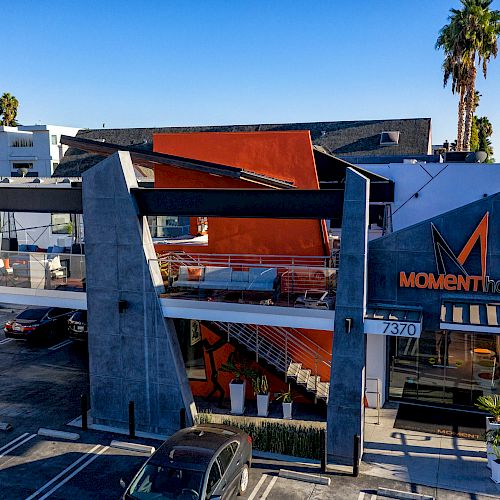 This image shows a modern building with a sign reading "MOMENT hotel," a parking lot in front, and palm trees in the background, under a clear sky.