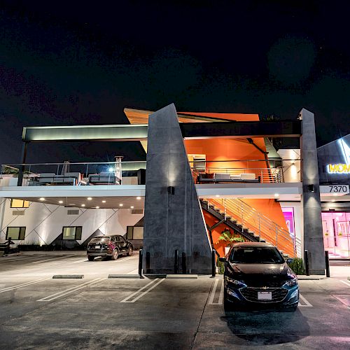 A modern two-story building at night with a brightly lit "M" logo sign, stairs, and parked cars in an open lot.