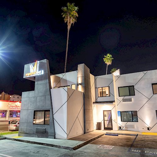 A modern, geometric building at night, with palm trees and streetlights. A colorful mural is visible on the nearby structure.