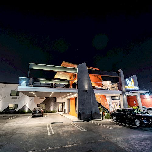 A modern two-story building at night with cars parked in the lot, featuring a prominent entrance and illuminated signage.