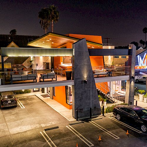 This image depicts a modern, elevated building with an illuminated sign at night, featuring a parking lot with cars below.