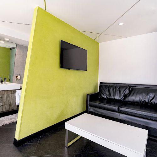 A modern room with a black couch, a TV on a green divider wall, a white coffee table, and a bathroom visible in the background.