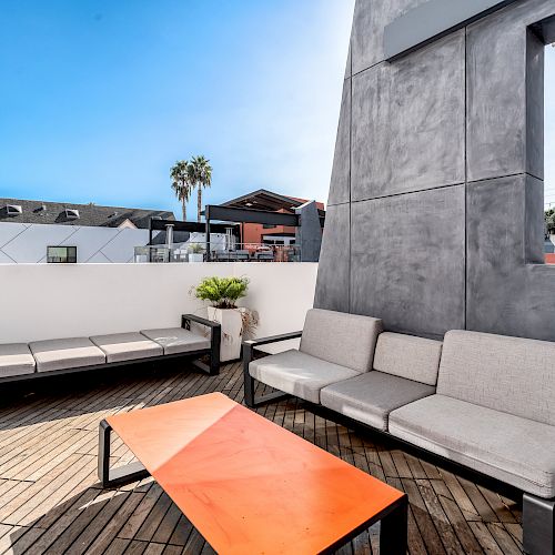 The image shows a modern outdoor patio with grey couches, a red table, and a view of buildings and palm trees under a clear blue sky.