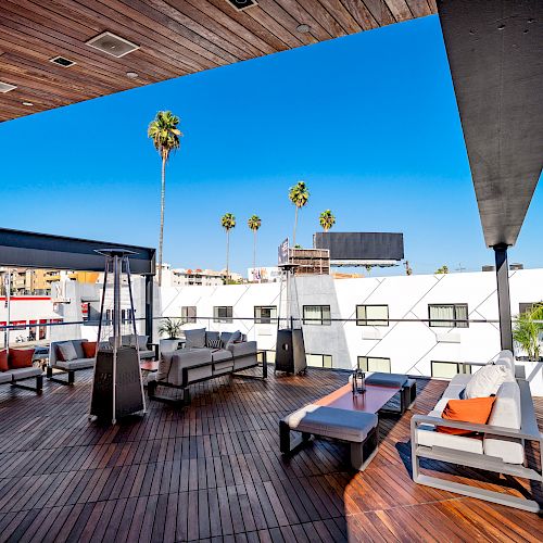 A rooftop lounge features modern seating, wooden flooring, and heaters. Palm trees and a clear blue sky are visible in the background.