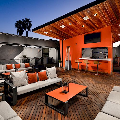 The image shows a modern outdoor lounge area with sofas, a bar with stools, and a wooden deck, featuring warm lighting and stylish decor.