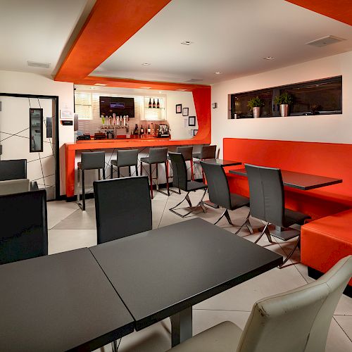 A modern bar with orange and black decor, featuring black chairs, orange cushioned benches, tables, and a stocked bar in the background.