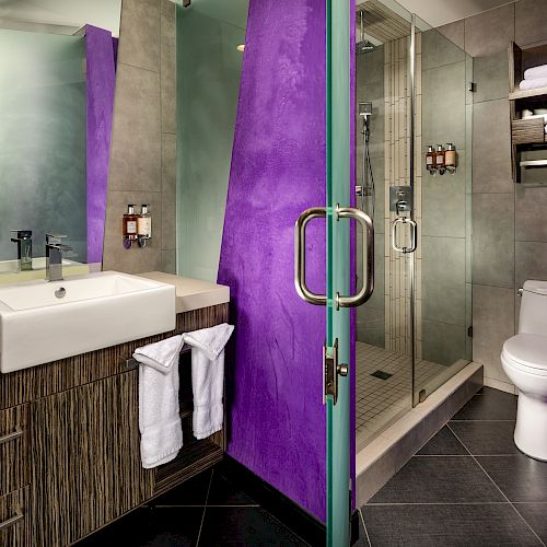 A modern bathroom with a sink, toilet, shower, purple accent walls, and shelving holding towels on a gray tiled wall ends the sentence.