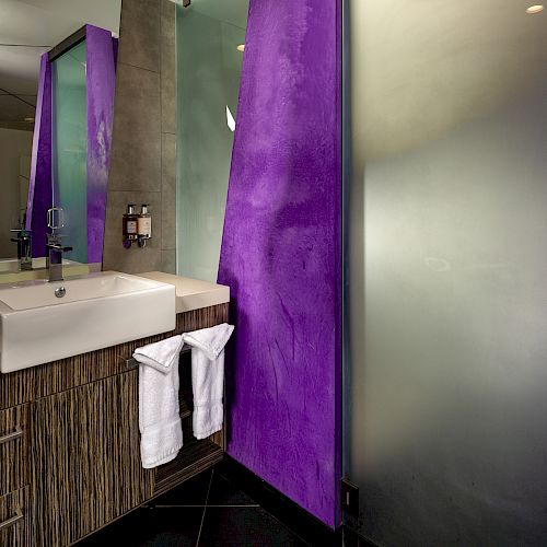 A modern bathroom with a sink, hand towels, a frosted glass door, and purple walls. The vanity has a drawer and a black floor can also be seen.