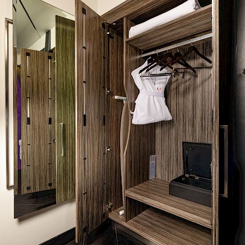 The image shows an open wooden wardrobe with shelves, a mirror, hangers, a neatly folded towel, a piece of clothing, and a small safe.