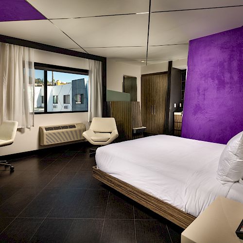 A modern hotel room with a purple accent wall, white bed, desk, chairs, window, and various furnishings. The overall design is contemporary.