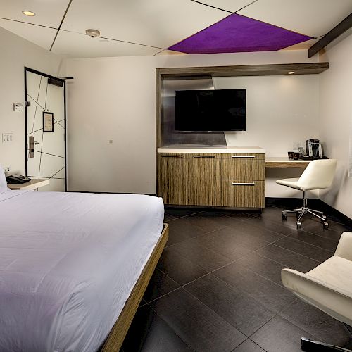 The image shows a modern hotel room with a bed, TV, desk, chair, window, and contemporary furnishings. The ceiling has a purple accent.