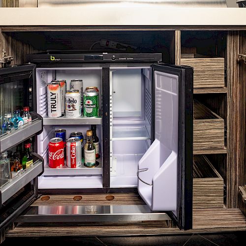 An open mini-fridge inside a cabinet contains various beverages, including sodas, beer, and water bottles. The fridge door has additional drinks.