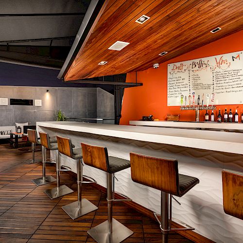 A modern bar with wooden stools, a white countertop, and an orange wall with a menu. There are couches in the background and a TV on the wall.