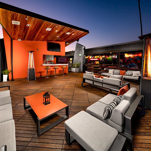 Stylish rooftop lounge with modern seating, wooden decor, ambient lighting, bar area, tables, and outdoor heaters. A relaxing spot for evening gatherings.