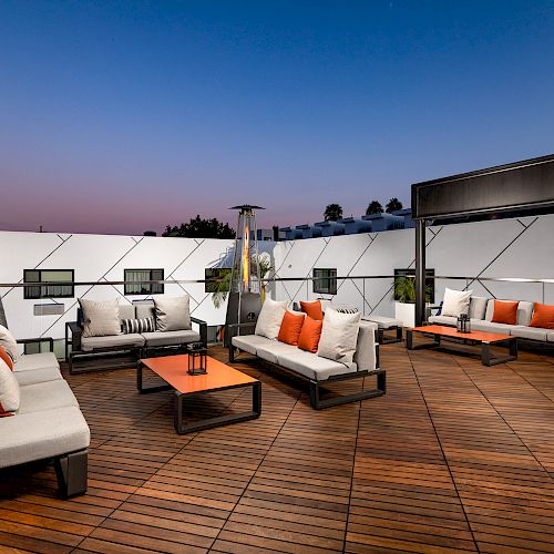 Modern rooftop lounge with white and orange cushioned seating, wooden decking, and a gas heater against a backdrop of a beautiful dusk sky.