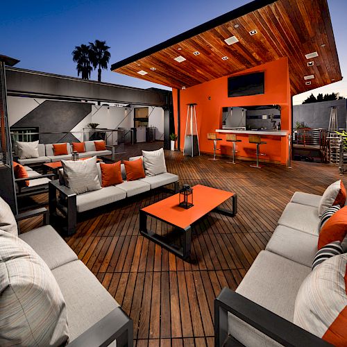 Modern outdoor lounge with cushioned sofas and chairs, wooden deck, orange accents, bar, stools, palm tree, and ambient lighting at dusk.